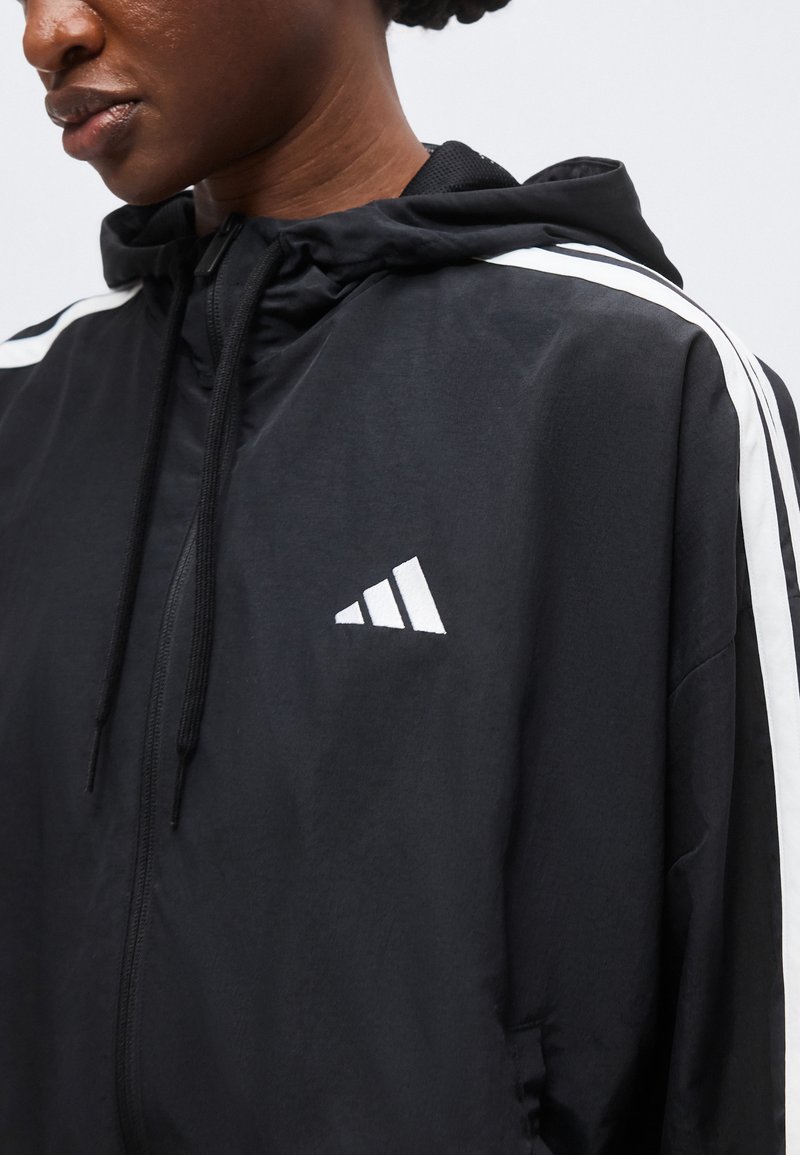 Persona che indossa una giacca nera con cappuccio e zip, con strisce bianche sulle spalle e un logo triangolare bianco sul petto.