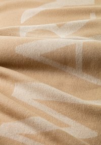 Beige stof met een zachte, gestructureerde oppervlakte, voorzien van een opvallend wit geometrisch patroon. Het materiaal lijkt warm en uitnodigend.