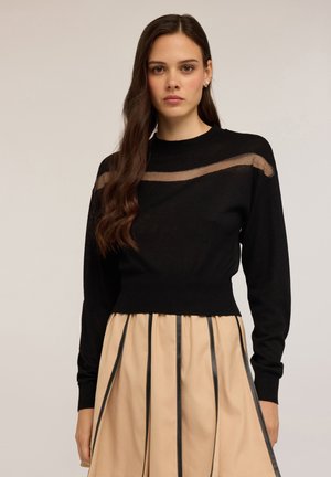 Pull noir en tricot avec une bande horizontale transparente sur la poitrine, taille cintrée et manches longues ; associé à une jupe plissée beige.