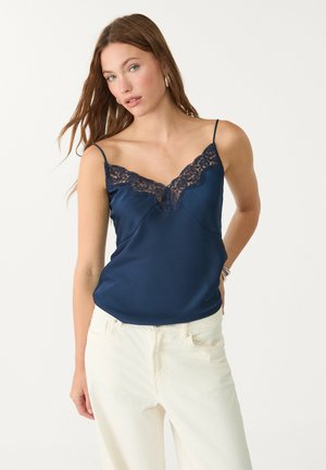 Camisole en satin marine avec une finition en dentelle au niveau du col. Dispose de fines bretelles et d'une coupe décontractée, complétée par un pantalon blanc taille haute.