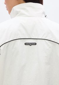 Λευκό μπουφάν αδιάβροχο με ψηλό γιακά, διακοσμημένο με μαύρη λεπτομέρεια και μαύρο λογότυπο "tommy jeans" στην πλάτη.