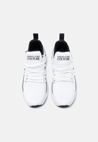 Άσπρα αθλητικά παπούτσια με μαύρες λεπτομέρειες, με επάνω μέρος από δίχτυ, επενδυμένο αυχένα και επίπεδα κορδόνια. Λογότυπο "Versace Jeans Couture" στη γλώσσα.