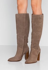 Botas de ante marrón hasta la rodilla con punta puntiaguda y tacón grueso. Cuentan con una cremallera lateral interior y una textura suave.