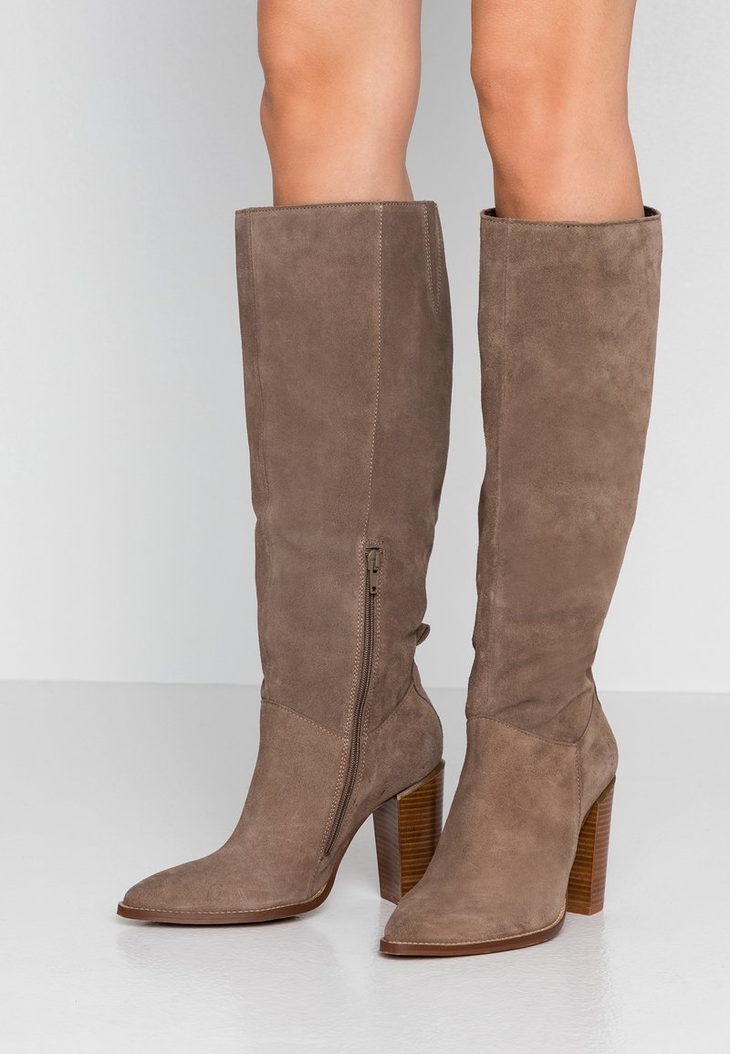 Botas de ante marrón hasta la rodilla con punta puntiaguda y tacón grueso. Cuentan con una cremallera lateral interior y una textura suave.