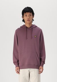 Sudadera morada con un bolsillo canguro, capucha con cordón y un pequeño logotipo amarillo. Tela suave y corte relajado. Llevada con pantalones de color claro.