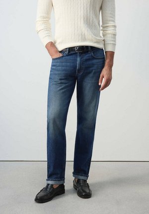 Homme portant un jean bleu foncé, une ceinture noire, un pull en maille torsadée crème et des mocassins en cuir noir, debout avec une main dans la poche.