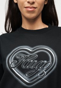 Νέος που φοράει μαύρο φούτερ με μια μεγάλη ασημί καρδιά και καλλιγραφικό κείμενο "Hugo" στο κέντρο του στήθους.