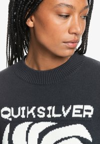 Tumehall villane sviiter ümmarguse kaelusega. Esipinnal on suur valge "Quiksilver" logo ja stiliseeritud lainekujundus.