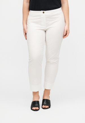 Γυναίκα που φοράει στενά λευκά παντελόνια μέχρι τον αστράγαλο με κούμπωμα με κουμπί, σε συνδυασμό με μαύρα σανδάλια τύπου slide με ανοιχτά δάχτυλα σε μονόχρωμο φόντο.