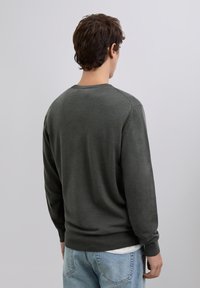 Dunkelgrüner Strickpullover mit Rundhalsausschnitt, langen Ärmeln und einer leicht texturierten Oberfläche, getragen über hellblauen Jeans.