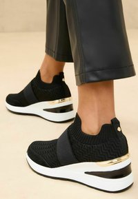 Baskets noires en tricot avec bande élastique, présentant un motif texturé, des accents dorés et une semelle compensée blanche avec un dessous noir.