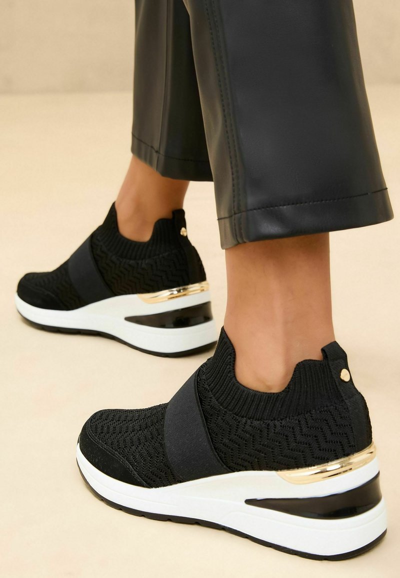 Baskets noires en tricot avec bande élastique, présentant un motif texturé, des accents dorés et une semelle compensée blanche avec un dessous noir.