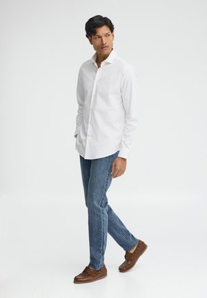 Uomo con camicia bianca con bottoni, jeans blu e mocassini di pelle marroni, in piedi con le mani nelle tasche davanti a uno sfondo bianco.