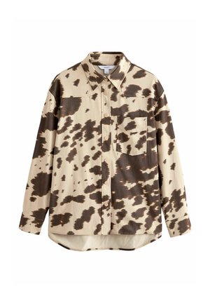 Camisa de manga larga en beige con estampado de vaca marrón. Presenta un cuello puntiagudo, cierre de botones y un único bolsillo en el pecho. Textura de tela suave.