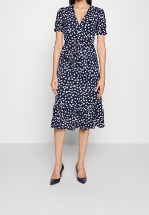 Vrouw draagt een marineblauwe wikkeljurk met witte bloemenprint, korte mouwen, een gestrikte taille en zwarte pumps met puntige neus, staand tegen een effen achtergrond.
