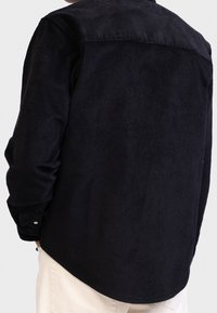 Persona con una camisa de corduroy negra de manga larga y puños abotonados, con pantalones beiges, mostrada de espaldas contra un fondo liso.