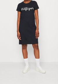Granatowa sukienka T-shirt z białym napisem "Hilfiger", krótkie rękawy, luźny krój i średniej długości dół. Nosi ją w białych butach typu slip-on.