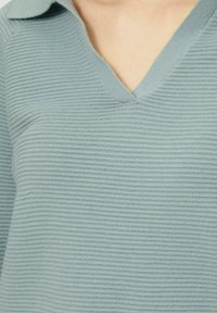 Hellblauer, gerippter Pullover mit V-Ausschnitt und Kragen. Der Stoff wirkt weich mit einer strukturierten Oberfläche und dezenten horizontalen Linien.