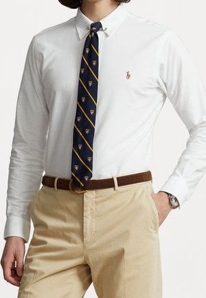 Homme portant une chemise blanche boutonnée avec logo, cravate à rayures bleu marine, pantalon beige, ceinture marron et montre-bracelet, une main dans la poche.
