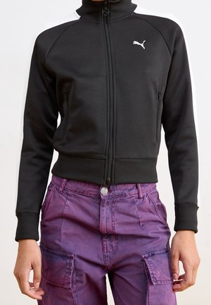 Gros plan d'une personne portant une veste zippée noire Puma avec des bandes blanches aux épaules et un pantalon cargo violet.