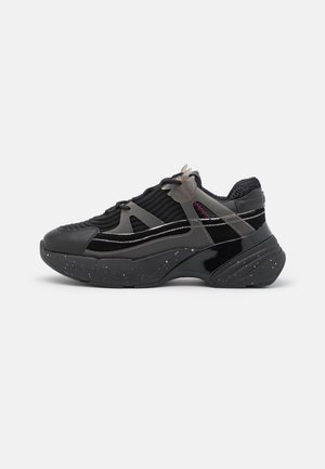 Deportiva negra voluminosa con parte superior de tela texturizada, suela gruesa con patrón moteado y correas de plástico gris a los lados y sobre los cordones.