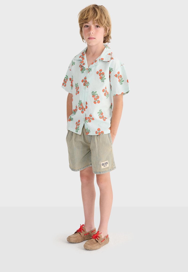 Enfant blond portant une chemise à manches courtes bleu clair avec un motif de fruits oranges, un short beige avec un patch, et des chaussures marron clair avec des lacets rouges, debout.