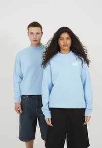 Lyseblå crewneck sweatshirt med ribbede mansjetter og halslinning. Modell som har på seg mørke denimshorts, en annen i svarte vide shorts.