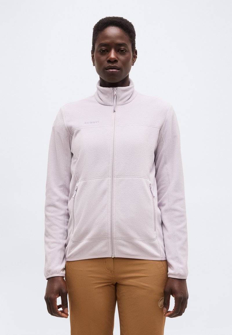 Άτομο που φοράει μωβ ανοιχτό φερμουάρ φλις μπουφάν και καφέ παντελόνι, στέκεται προς τα εμπρός μπροστά σε μια απλή λευκή φόντο.