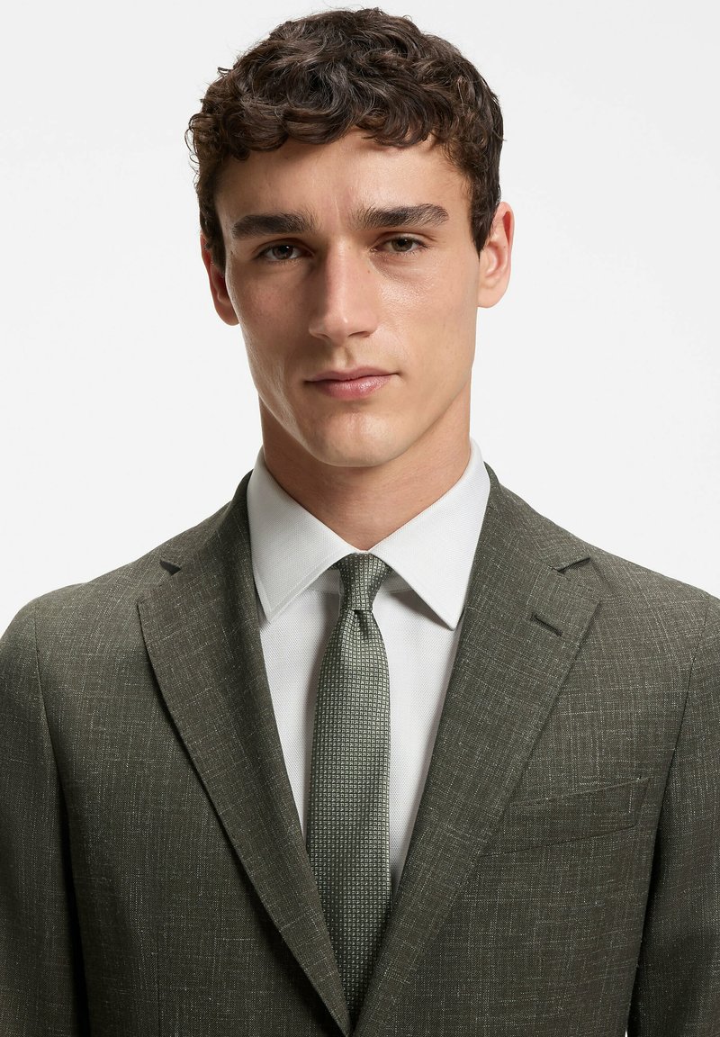 Jeune homme aux cheveux bruns bouclés portant un costume texturé vert olive, une chemise blanche et une cravate à motifs, fait face à l’avant avec une expression neutre.