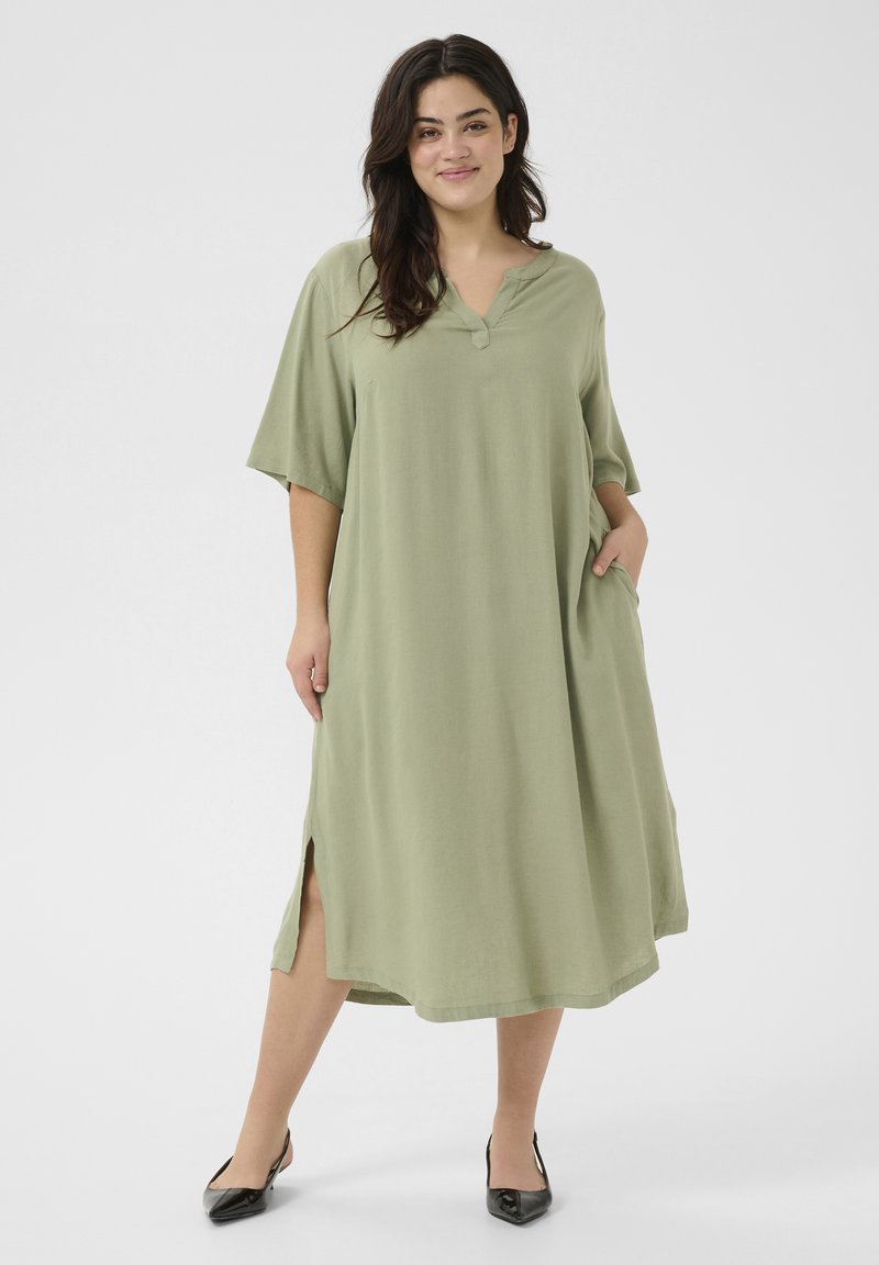 Vestido verde hasta la rodilla con mangas cortas, escote en V y aberturas laterales. Tejido suave con un corte holgado. La modelo lleva zapatos planos negros.