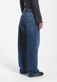 Szerokie niebieskie dżinsy z wyblakłym wykończeniem, z wysokim stanem, dwoma tylnymi kieszeniami oraz detalami w postaci szwów wzdłuż dołów nogawek.