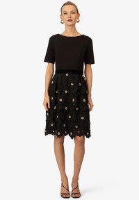 Robe noire avec un corsage ajusté et des manches courtes, dotée d'une jupe en dentelle florale ornée de touches dorées et d'un ourlet en forme de scallop.