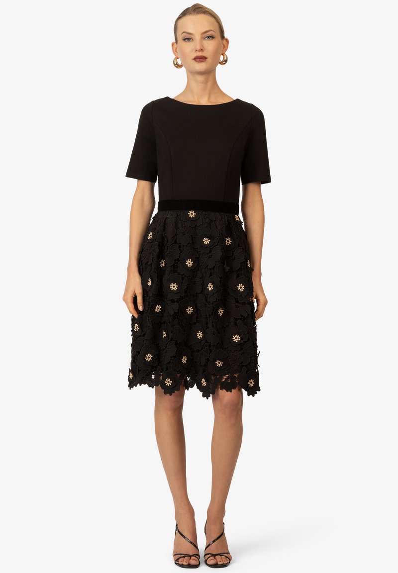 Robe noire avec un corsage ajusté et des manches courtes, dotée d'une jupe en dentelle florale ornée de touches dorées et d'un ourlet en forme de scallop.