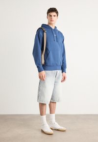 Hoodie in cotone blu con cordini e tasca anteriore, abbinato a pantaloni shorts in denim chiaro. Sneakers bianche e calzini bianchi di lunghezza media completano il look.