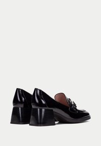 Zapatos de tacón de charol negro con un tacón grueso y estriado y un diseño de punta redondeada y suave. Con un detalle decorativo de lazo en la parte delantera.