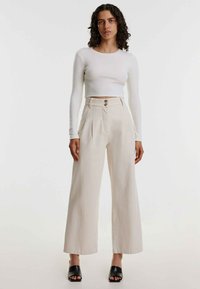 Beige weite Hose mit Falten und zwei vorderen Knöpfen, kombiniert mit einem enganliegenden weißen Langarmoberteil und schwarzen offenen Mules.