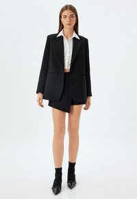Schwarze Blazer mit Ausschlagrevers, kombiniert mit einem knappen weißen Hemd und einem schwarzen Minirock mit asymmetrischem Saum. Schwarze spitze Absätze.