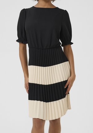 Donna che indossa un vestito nero con maniche a sbuffo e una gonna plissettata con ampie strisce orizzontali nere e crema.