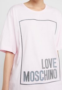 Růžové tričko s krátkým rukávem s černým ohraničením a nápisem "LOVE MOSCHINO" vytištěným texturovaným písmem vlevo dole. Hladký bavlněný materiál.
