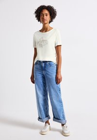 T-shirt créme à manches courtes avec imprimé graphique, associé à un jean large bleu clair et des baskets crème, avec les ourlets du jean retroussés.