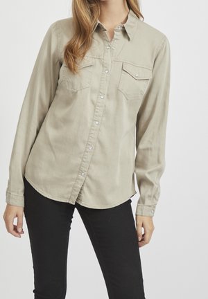 Femme portant une chemise beige clair à manches longues avec poches poitrine et pantalon noir ajusté, debout devant un fond uni.