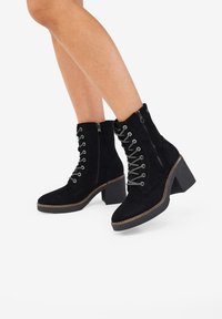 Stivaletti neri in suede con punta tonda, lacci sulla parte anteriore, zip laterale e tacco grosso. Presentano cuciture a contrasto e una suola testurizzata.