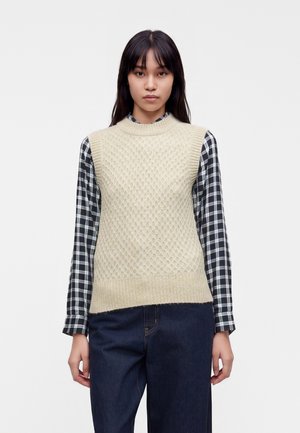 Femme portant un pull sans manches en maille crème par-dessus une chemise à carreaux noir et blanc à manches longues et un jean bleu foncé, debout devant un fond blanc.