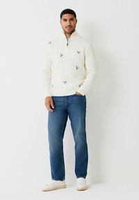 Balta adīta džemperis ar puszips apkakli, rotāts ar krāsainiem slēpošanas motīviem, apvienots ar ziliem džinsiem un baltiem sporta apaviem.