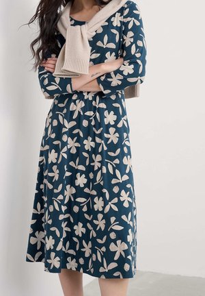 Kvinde iført en blå midikjole med beige blomsterprint, en beige sweater hængende over skuldrene, arme krydset, stående mod en ensfarvet væg.