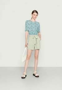 Top floral de manga curta em azul e verde, combinado com calções de cintura alta em verde claro com um laço. Modelo segura uma bolsa branca.