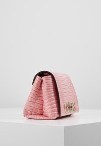 Lyserød krokodillepræget håndtaske med en struktureret silhuet, guld hardware-logo og kontrasterende sort trim. Teksturerede overfladedetaljer synlige.