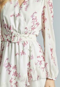 Abito floreale con maniche lunghe, dotato di scollo a V e vita cintata. Tessuto bianco decorato con motivi di fiori rosa e rifiniture a volant.