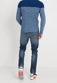 Mann in einem farbigen blauen Blockpullover, abgetragenen blauen Jeans und weißen Sneakers mit roten und schwarzen Details, der vor einem schlichten weißen Hintergrund weggeht.