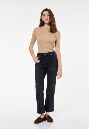 Jonge vrouw met golvend bruin haar draagt een beige ribgebreide top met korte mouwen, zwarte high-waist spijkerbroek en bruine mocassins, staand tegen een witte achtergrond.